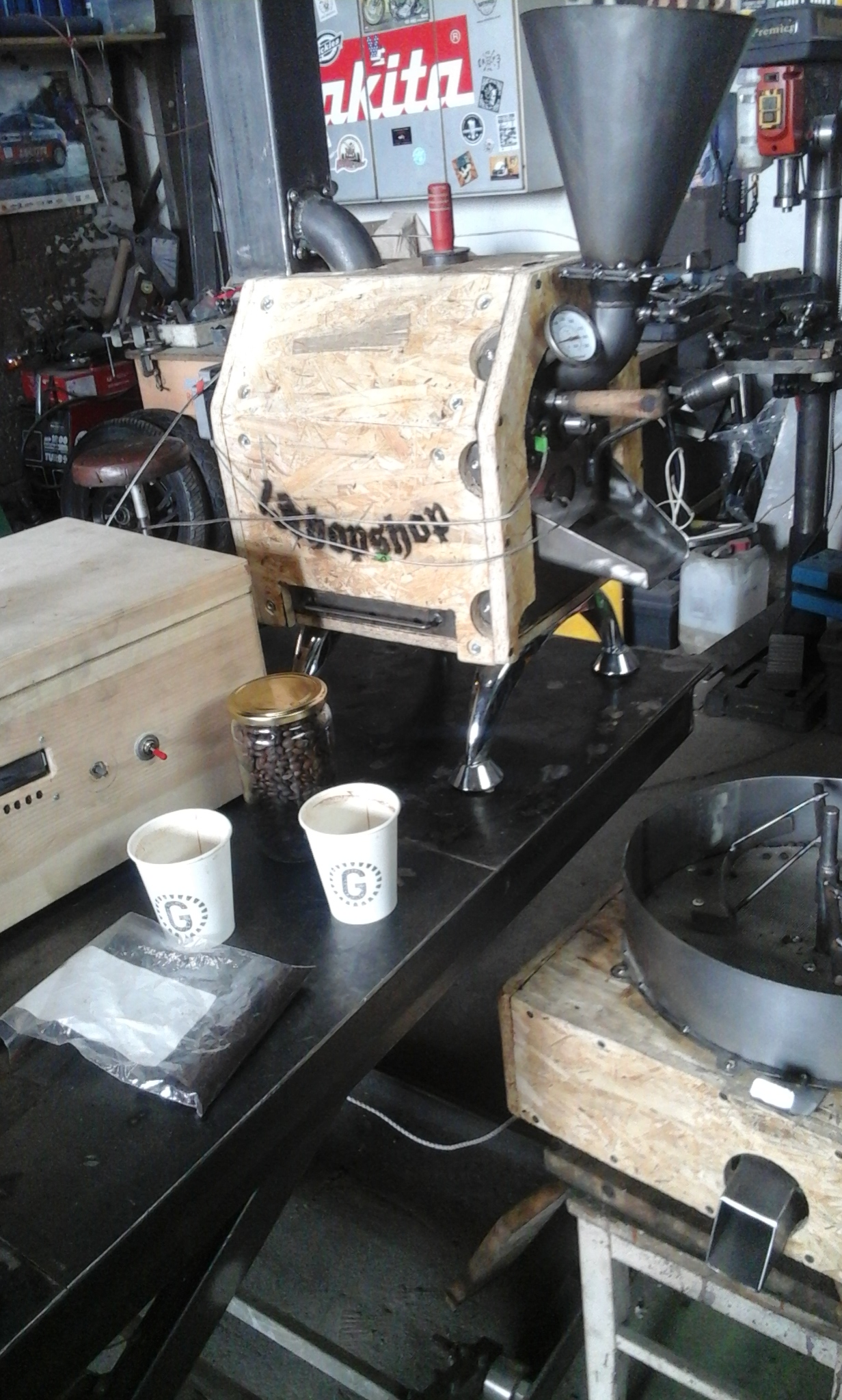 Ręcznie robiona palarnia kawy z obudową z płyty OSB, widoczne ziarna kawy, kubki i paczka kawy na warsztatowym stole.