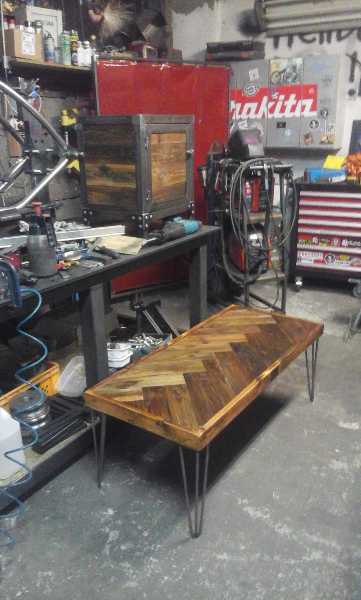 Stolik kawowy z drewnianym blatem ułożonym w jodełkę i metalowymi nogami, obok szafka z metalową ramą i drewnianymi drzwiczkami, w tle warsztat z narzędziami i sprzętem spawalniczym.