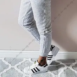 Osoba w szarych spodniach dresowych i białych sneakersach z czarnymi paskami, stojąca na wzorzystym dywanie w pomieszczeniu z jasną ścianą.