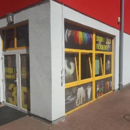 Fasada budynku z żółtymi ramami okiennymi i drzwiowymi, na których umieszczono grafiki prezentujące przykładowe wydruki 3D i napisy 'Druk i Filamenty 3D', z widocznymi modelami wydrukowanymi w 3D...