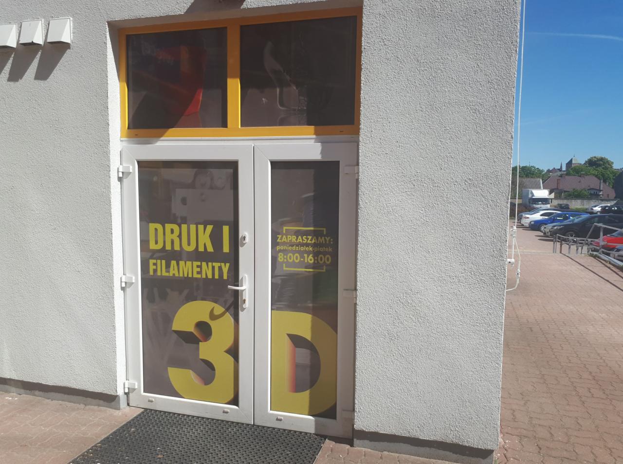Wejście do punktu usługowego z naklejonym na drzwiach napisem 'Druk i filamenty' oraz '3D', z informacją o godzinach otwarcia od poniedziałku do piątku.