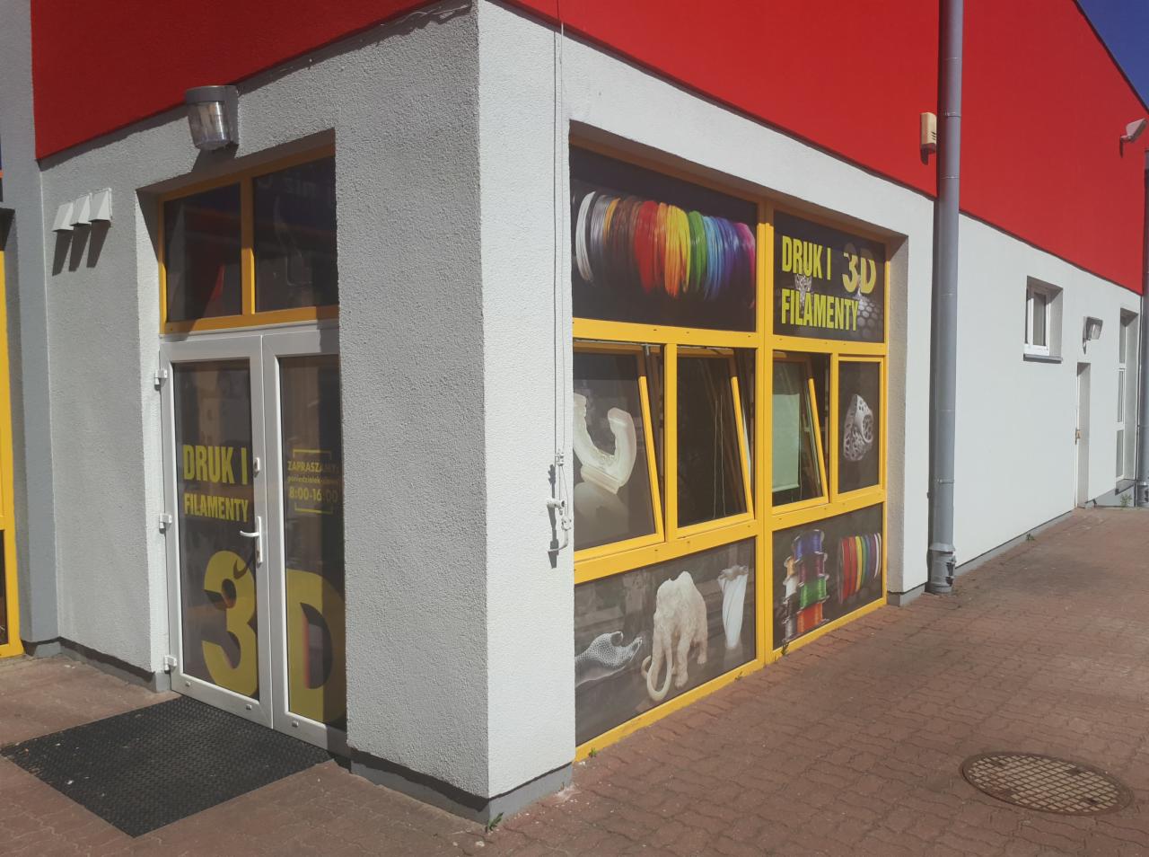 Fasada budynku z żółtymi ramami okiennymi i drzwiowymi, na których umieszczono grafiki prezentujące przykładowe wydruki 3D i napisy 'Druk i Filamenty 3D', z widocznymi modelami wydrukowanymi w 3D...
