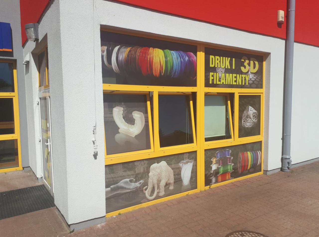 Fasada budynku z oknami oklejonymi folią prezentującą wydruki 3D i kolorowe filamenty, z napisem 'DRUKI 3D FILAMENTY'.