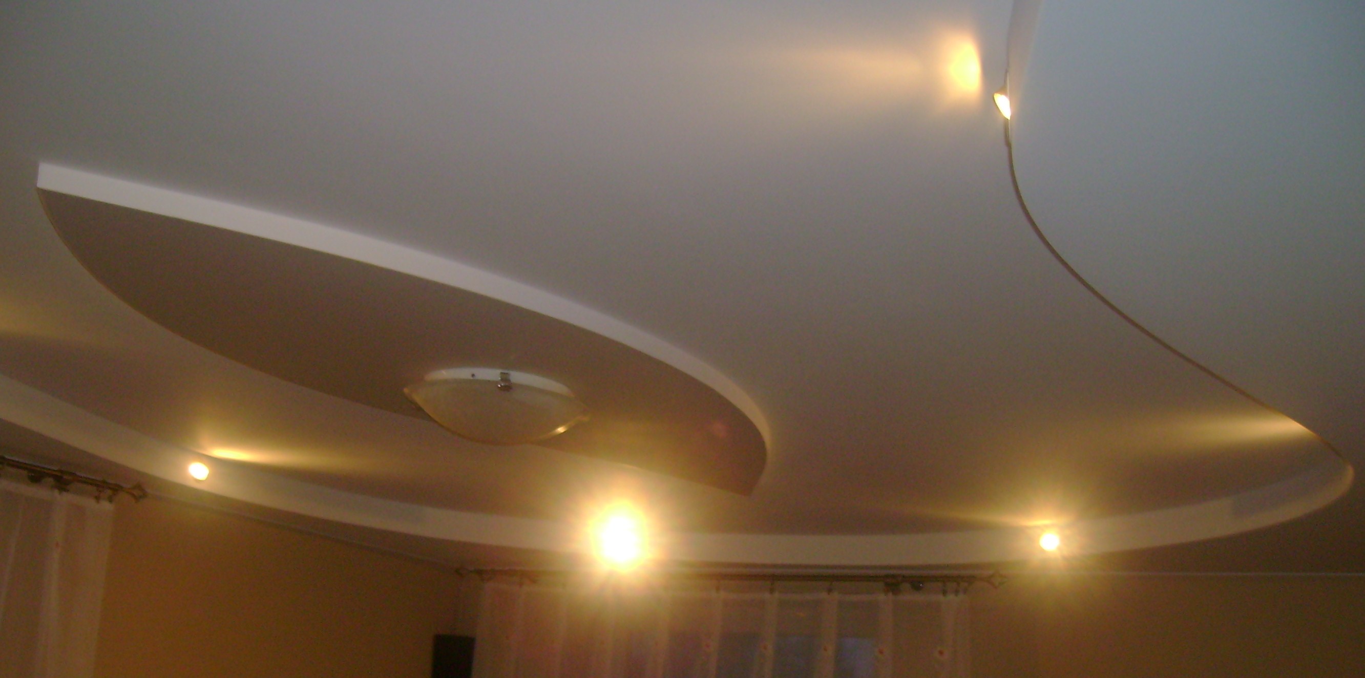 Nowoczesny sufit napinany z oświetleniem punktowym i centralnym w Łodzi, widok z dołu, dekoracyjne wykończenie w kształcie łuku.