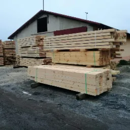 Stosy świeżo przetartego drewna budowlanego, zabezpieczone zieloną taśmą, ułożone na betonowych podporach przed budynkiem gospodarczym w pochmurny dzień.