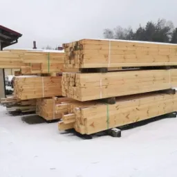 Stosy świeżo przetartego drewna budowlanego, zabezpieczone taśmami, składowane na śniegu przed budynkiem.