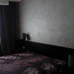 Spektor Plus - Ściana w sypialni z dekoracyjnym malowaniem w odcieniach szarości, z widoczną fakturą, nad czarnym zagłówkiem łóżka z pościelą w kwiaty, oświetlona przez wiszące lampy.