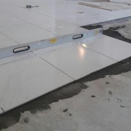 Spektor Plus - Układanie białych, marmurkowych płytek podłogowych z użyciem poziomicy i krzyżyków dystansowych, widoczna świeża zaprawa.