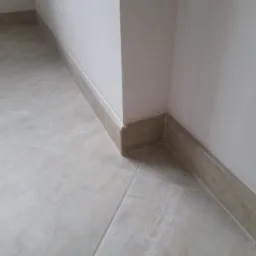 Wykończony narożnik pomieszczenia z beżowymi płytkami podłogowymi i listwami przypodłogowymi, widoczne połączenie płytek i listew przy ścianie.