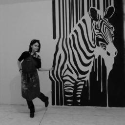 Spektor Plus - Kobieta w ubrudzonym farbą fartuchu pozuje obok czarno-białego muralu zebry z efektem spływającej farby i elementami kodu kreskowego na ścianie.