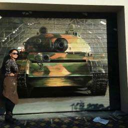 Spektor Plus - Kobieta w okularach i brązowym fartuchu stoi obok garażowych drzwi segmentowych z namalowanym kamuflażowym czołgiem na tle hali.