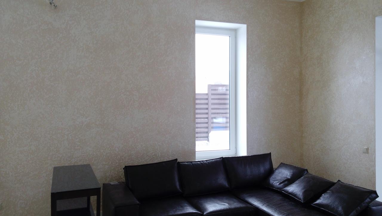 Beżowa ściana z dekoracyjną fakturą, wąskie okno z widokiem na budynek, czarna skórzana sofa narożna i mały stolik.