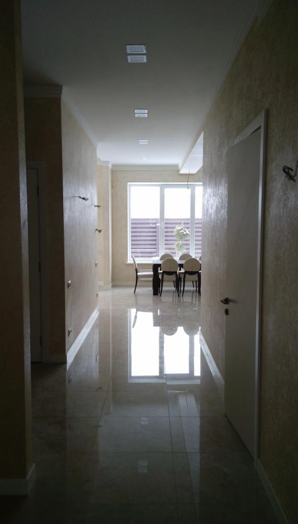 Korytarz w jasnych barwach z błyszczącą podłogą, widok na jadalnię z oknem i zasłoniętymi żaluzjami, ściany z dekoracyjną fakturą, białe drzwi po prawej stronie.