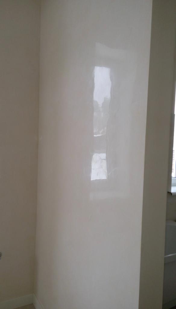 Ściana w rogu pokoju z wykończeniem w jasnym, połyskującym odcieniu, odbijająca okno, widoczne delikatne niedoskonałości faktury.