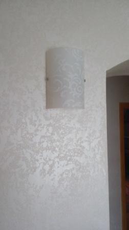 Ściana z dekoracyjną strukturą w odcieniach bieli, oświetlona przez kinkiet z delikatnym wzorem, widoczny fragment futryny drzwi w tle.