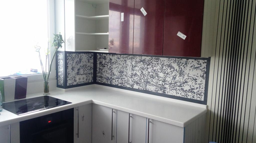 Nowoczesna kuchnia z białymi szafkami, blatem i panelem ściennym z abstrakcyjnym wzorem w odcieniach szarości, czarna płyta indukcyjna i dekoracyjne bambusy w wazonie na blacie.