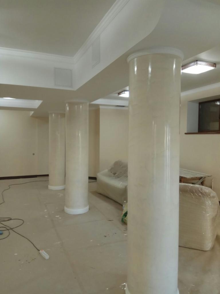 Wnętrze z kolumnami dekoracyjnymi wykończonymi w jasnym marmurkowym wzorze, widoczne fragmenty sufitu z listwami ozdobnymi oraz meble zabezpieczone folią ochronną.