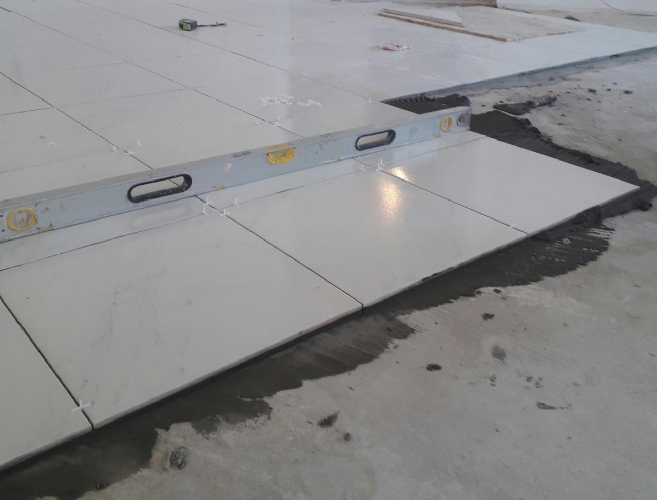 Układanie białych, marmurkowych płytek podłogowych z użyciem poziomicy i krzyżyków dystansowych, widoczna świeża zaprawa.