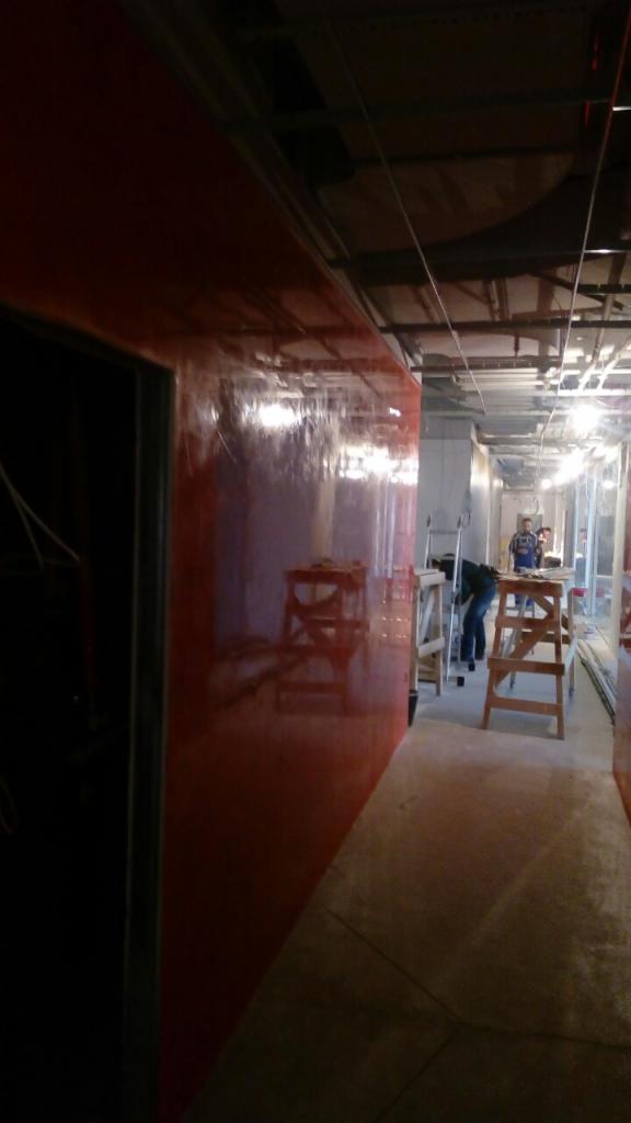 Korytarz w trakcie prac wykończeniowych, ściana pokryta czerwoną, błyszczącą farbą, widoczne drabiny i pracownicy w oddali.