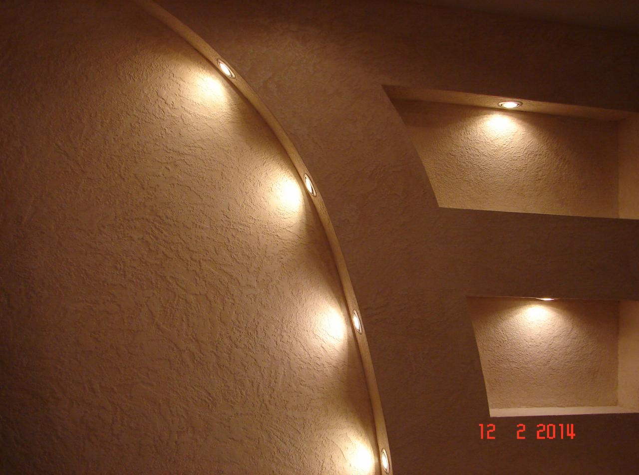 Ściana z dekoracyjną fakturą, wnękami i oświetleniem punktowym, tworząca łuk i półki podświetlone ciepłym światłem.