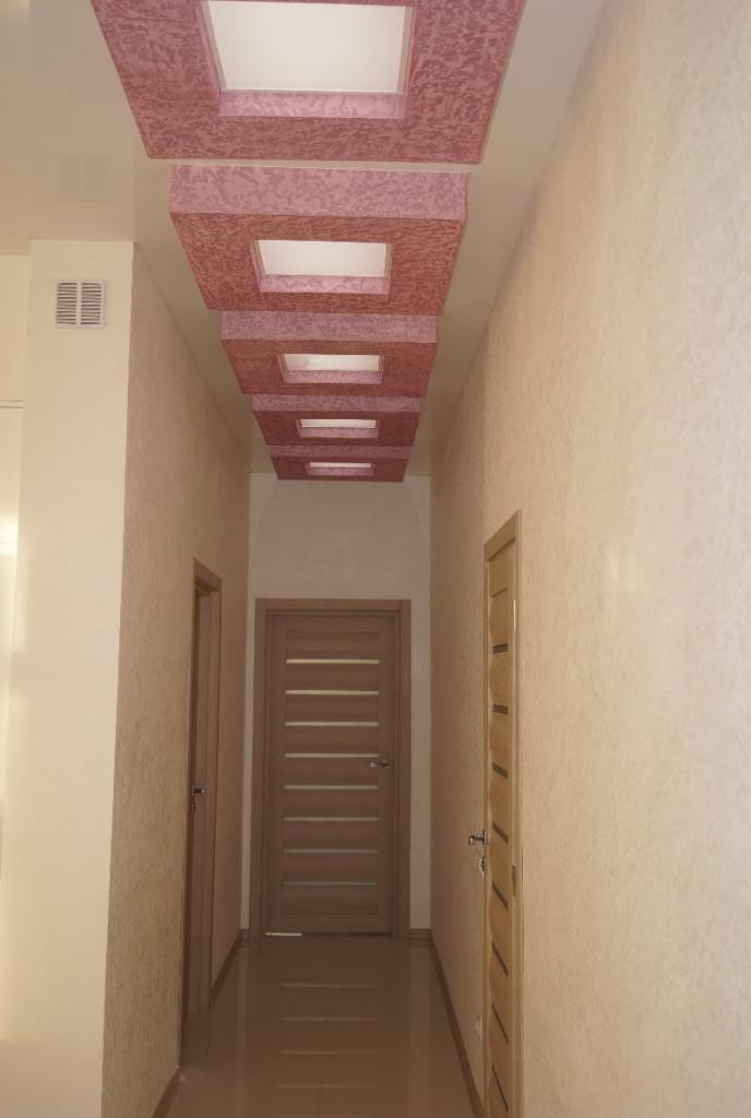 Korytarz z dekoracyjnym, kaskadowym sufitem podwieszanym w odcieniach różu, z wbudowanym oświetleniem i dwiema drewnianymi drzwiami po bokach.