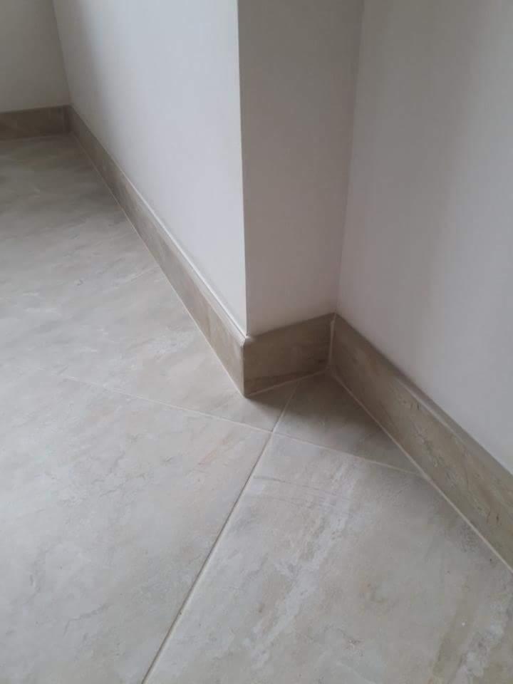 Wykończony narożnik pomieszczenia z beżowymi płytkami podłogowymi i listwami przypodłogowymi, widoczne połączenie płytek i listew przy ścianie.