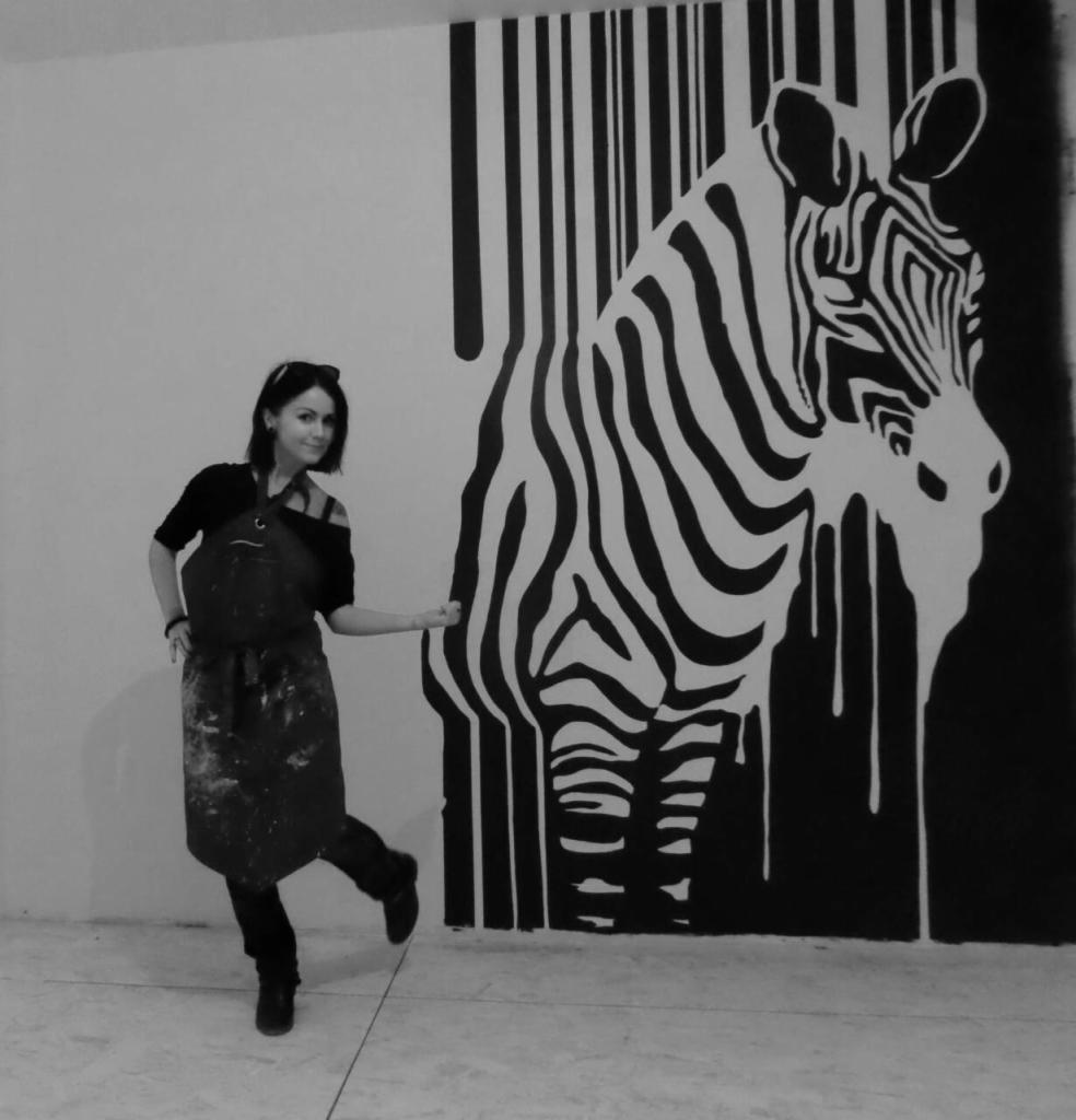 Kobieta w ubrudzonym farbą fartuchu pozuje obok czarno-białego muralu zebry z efektem spływającej farby i elementami kodu kreskowego na ścianie.