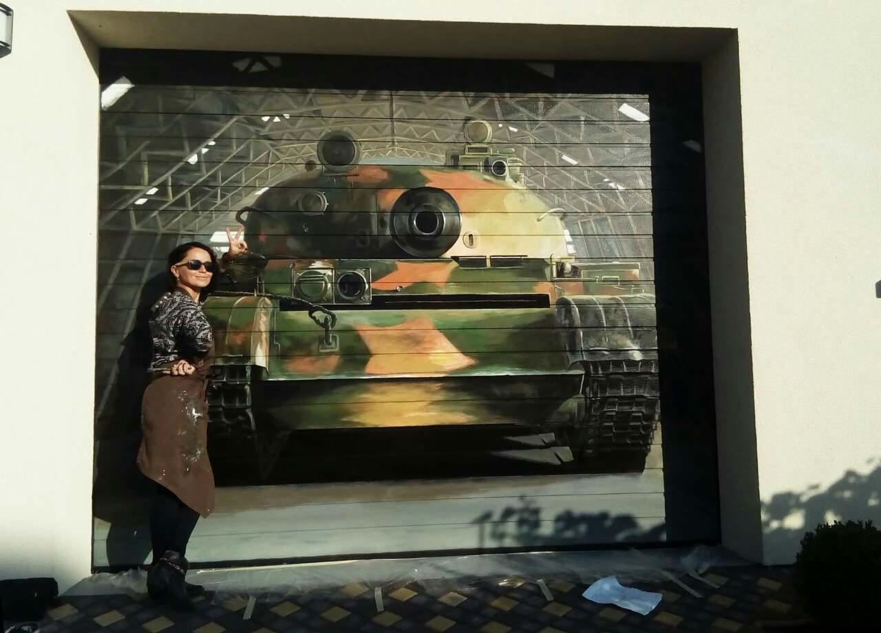 Kobieta w okularach i brązowym fartuchu stoi obok garażowych drzwi segmentowych z namalowanym kamuflażowym czołgiem na tle hali.