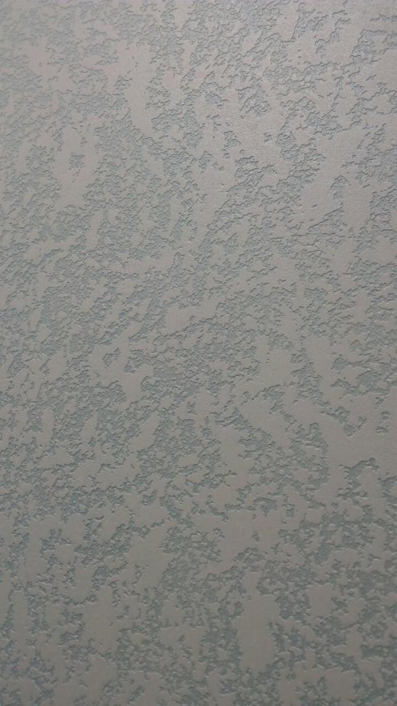 Szara ściana z wyraźną, nieregularną fakturą, przypominającą drobne grudki lub krople, tworzące wzór na powierzchni.