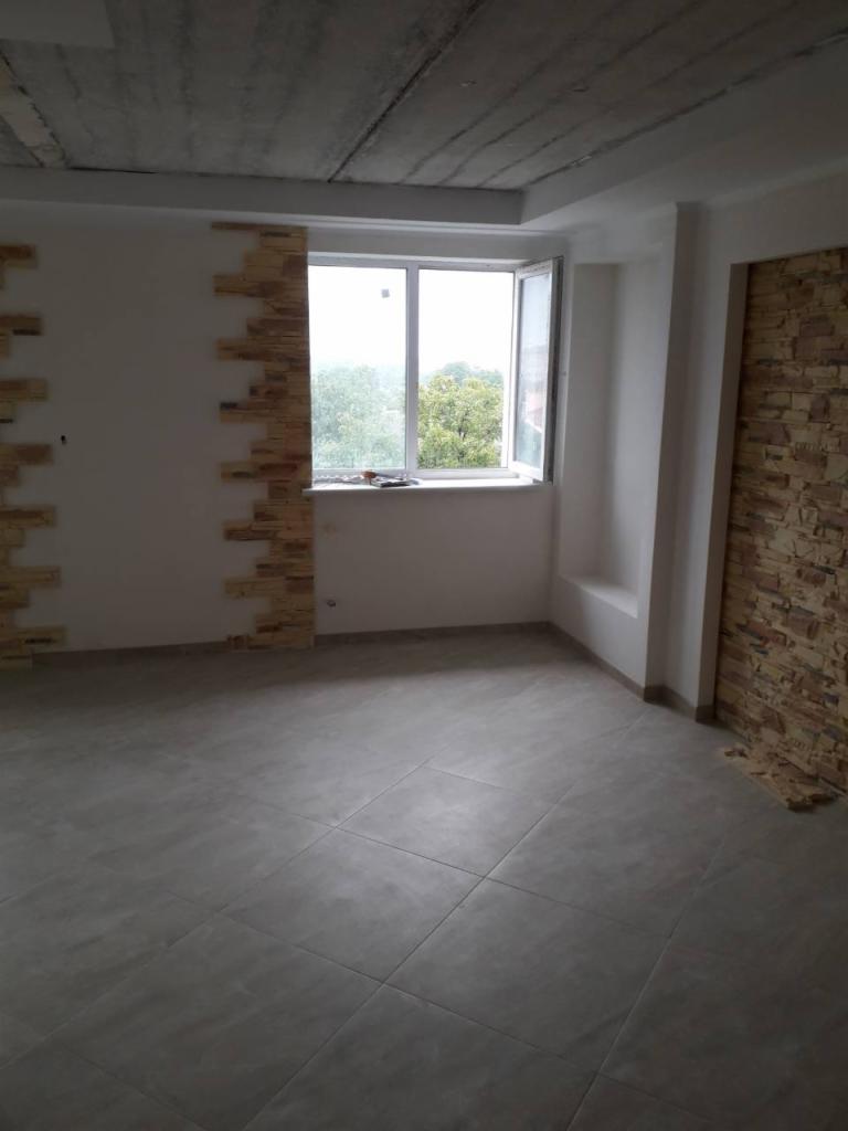 Wnętrze pokoju w trakcie wykańczania: częściowo wyłożona kamieniem dekoracyjnym ściana, surowy betonowy sufit, szare płytki podłogowe, białe okno z widokiem na zieleń.