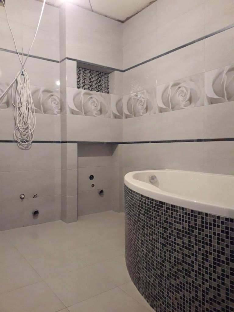 Wanna z mozaiką w łazience z białymi płytkami z motywem róży, widoczne przygotowane otwory na armaturę, przewody zwisające ze ściany.