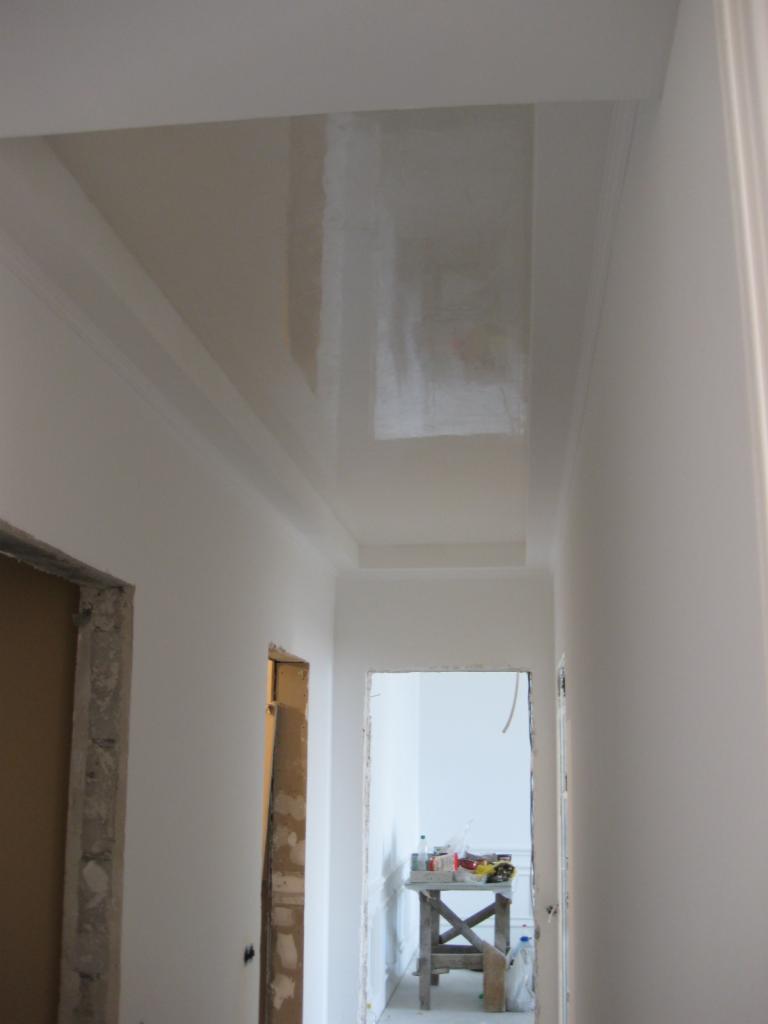 Korytarz w trakcie prac wykończeniowych, widoczny sufit z elementami szpachlowania, ściany po malowaniu na biało, ościeżnice drzwiowe w trakcie montażu.