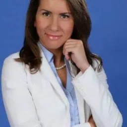 Portret uśmiechniętej kobiety w białym żakiecie, błękitnej koszuli i perłach, z ręką podpartą pod brodą na tle niebieskiego tła.