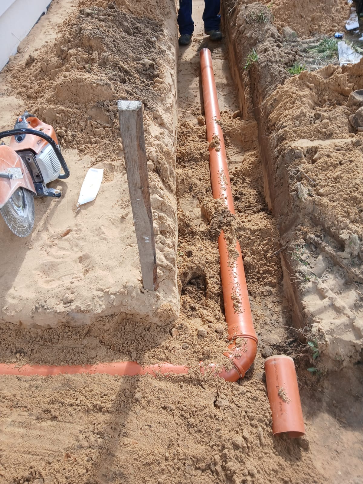 Pomarańczowe rury kanalizacyjne ułożone w wykopie, widoczna piła do cięcia rur i drewniany słupek. Częściowo widoczne nogi osoby nad wykopem.