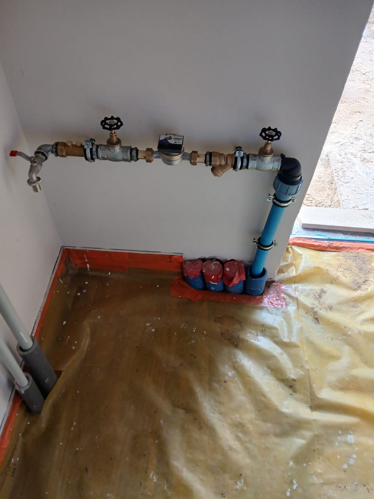 Kompletne przyłącze wodno-kanalizacyjne z licznikiem, zaworami i rurami, zamontowane przy ścianie wewnątrz budynku, z widocznym zabezpieczeniem podłoża folią.