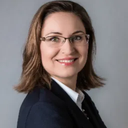 Portret uśmiechniętej kobiety w okularach i granatowym żakiecie na szarym tle.