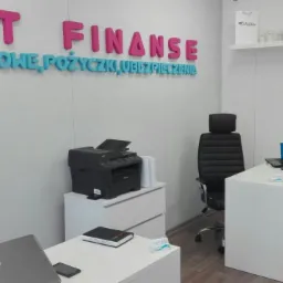 Wnętrze biura z napisem na ścianie 'TBet Finanse', biurko z laptopem i niebieskim kubkiem, drukarka na szafce z szufladami.