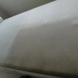 Fragment białego materaca z widocznym efektem czyszczenia, gdzie jedna część jest wyraźnie jaśniejsza od drugiej, obok wezgłowie z pikowaniem i kryształkami.