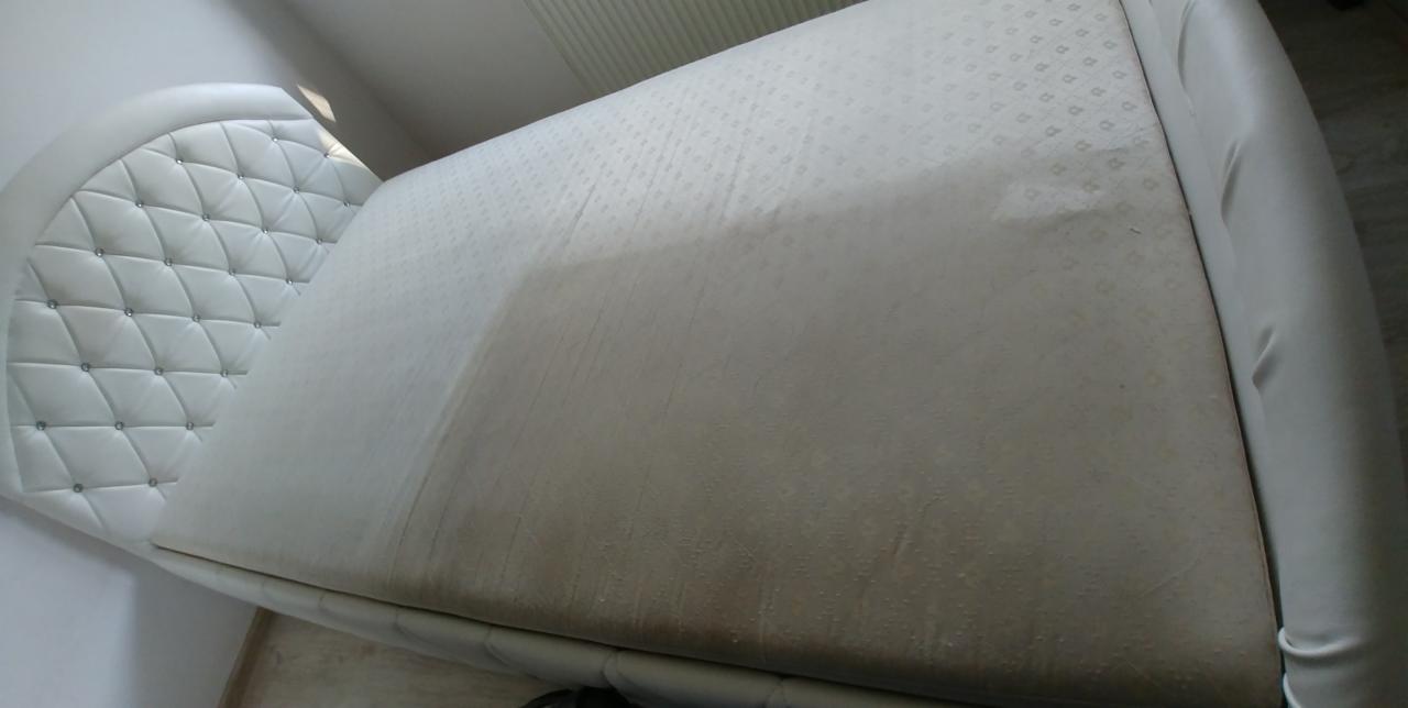 Fragment białego materaca z widocznym efektem czyszczenia, gdzie jedna część jest wyraźnie jaśniejsza od drugiej, obok wezgłowie z pikowaniem i kryształkami.