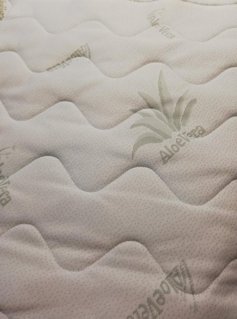 Jasny, pikowany pokrowiec na materac z delikatnym wzorem Aloe Vera, widoczne regularne przeszycia tworzące fale.
