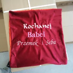 Bordowa poduszka z haftowanym napisem 'Kochanej Babci Przemek i Seba', leżąca na kartonowym pudełku.