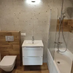 Nowoczesna łazienka z beżowymi płytkami imitującymi kamień, drewnopodobnymi panelami za toaletą, białą umywalką z szafką i wanną z parawanem.