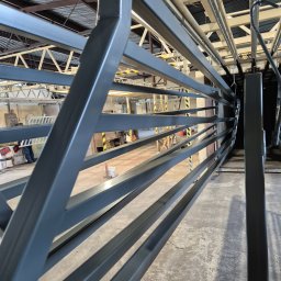 PKB TONDEL sc Paweł Robert Lilia Tondel - Szara, metalowa konstrukcja z poziomymi prętami w hali produkcyjnej, widoczne elementy suwnicy i podłoga z widocznymi śladami użytkowania.