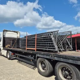 Ciężarówka przewozi metalowe konstrukcje, prawdopodobnie elementy ogrodzeń lub balustrad, zabezpieczone pomarańczowymi pasami, na tle błękitnego nieba z chmurami.