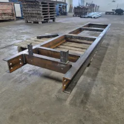 Metalowa rama konstrukcyjna z profili stalowych na palecie w hali produkcyjnej, widoczne spawane łączenia i otwory montażowe, w tle inne konstrukcje i materiały.