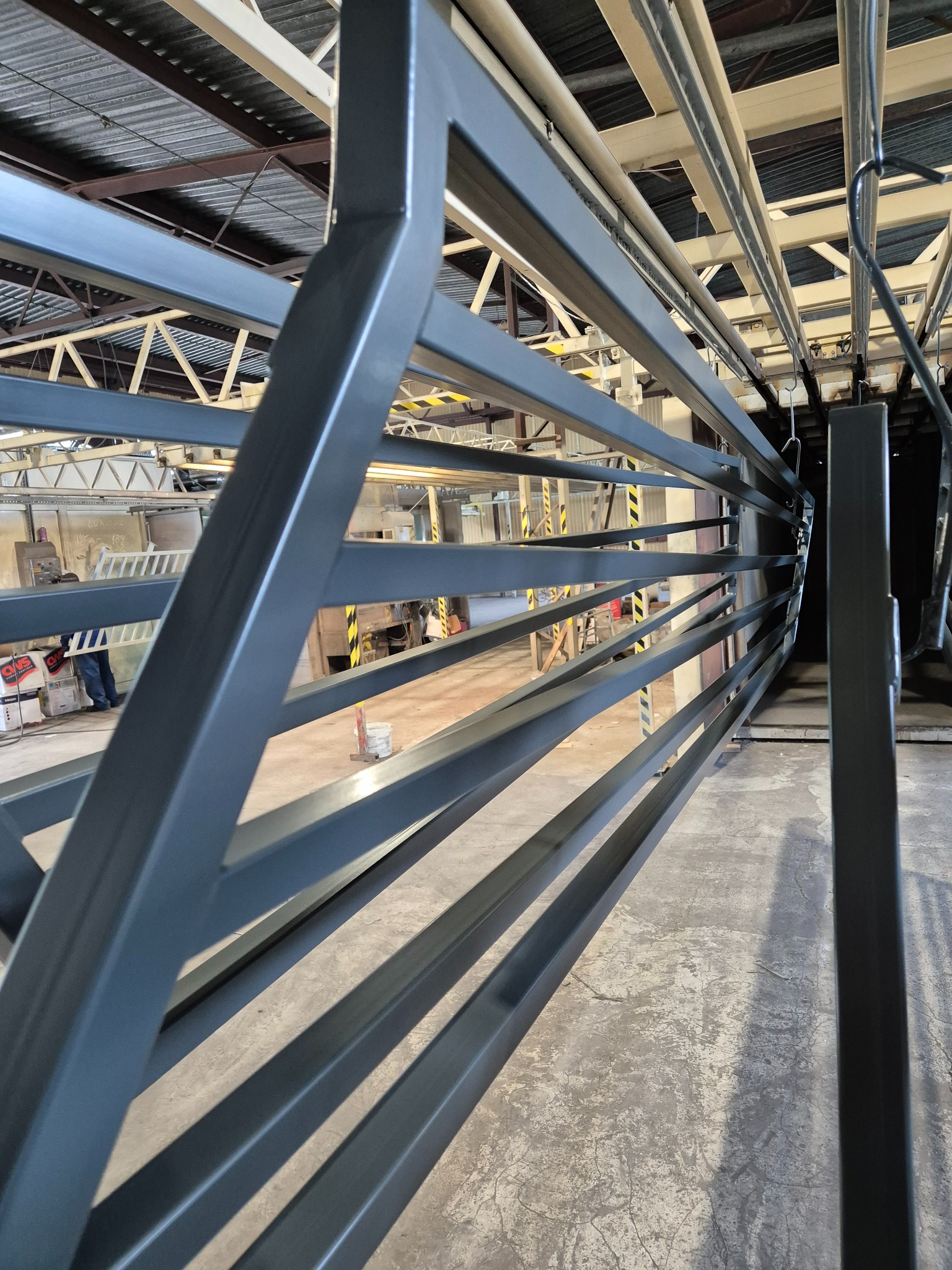 Szara, metalowa konstrukcja z poziomymi prętami w hali produkcyjnej, widoczne elementy suwnicy i podłoga z widocznymi śladami użytkowania.