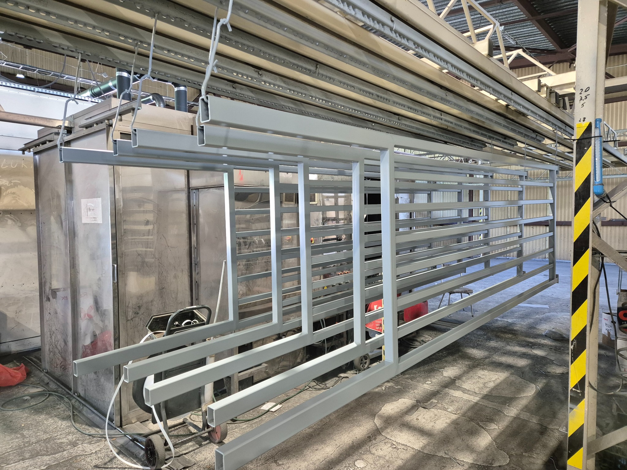 Elementy metalowe konstrukcji, prawdopodobnie ogrodzenia, zawieszone na łańcuchach w hali produkcyjnej, przygotowane do malowania proszkowego lub po nim, z widocznym piecem do malowania w tle.