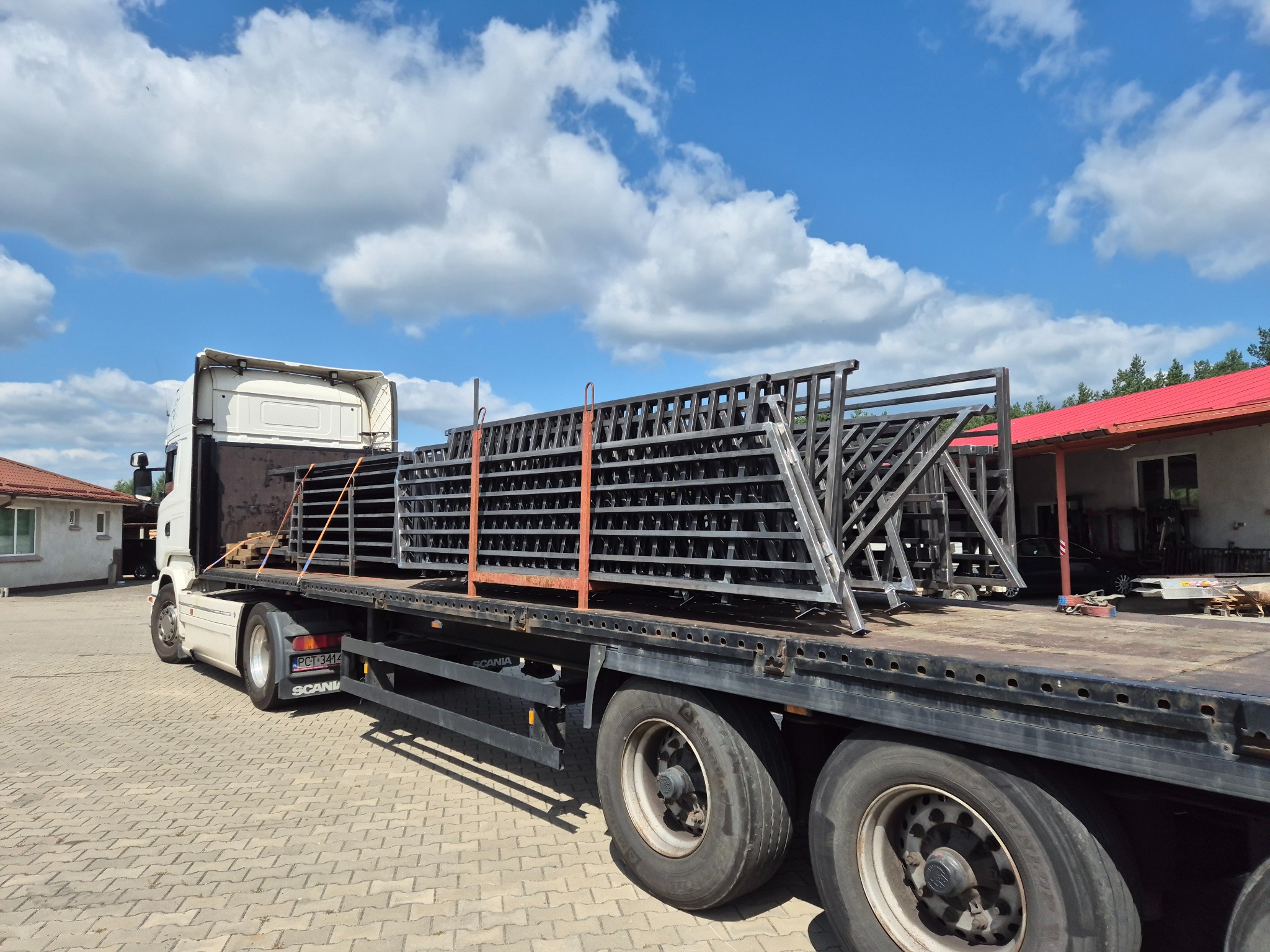 Ciężarówka przewozi metalowe konstrukcje, prawdopodobnie elementy ogrodzeń lub balustrad, zabezpieczone pomarańczowymi pasami, na tle błękitnego nieba z chmurami.