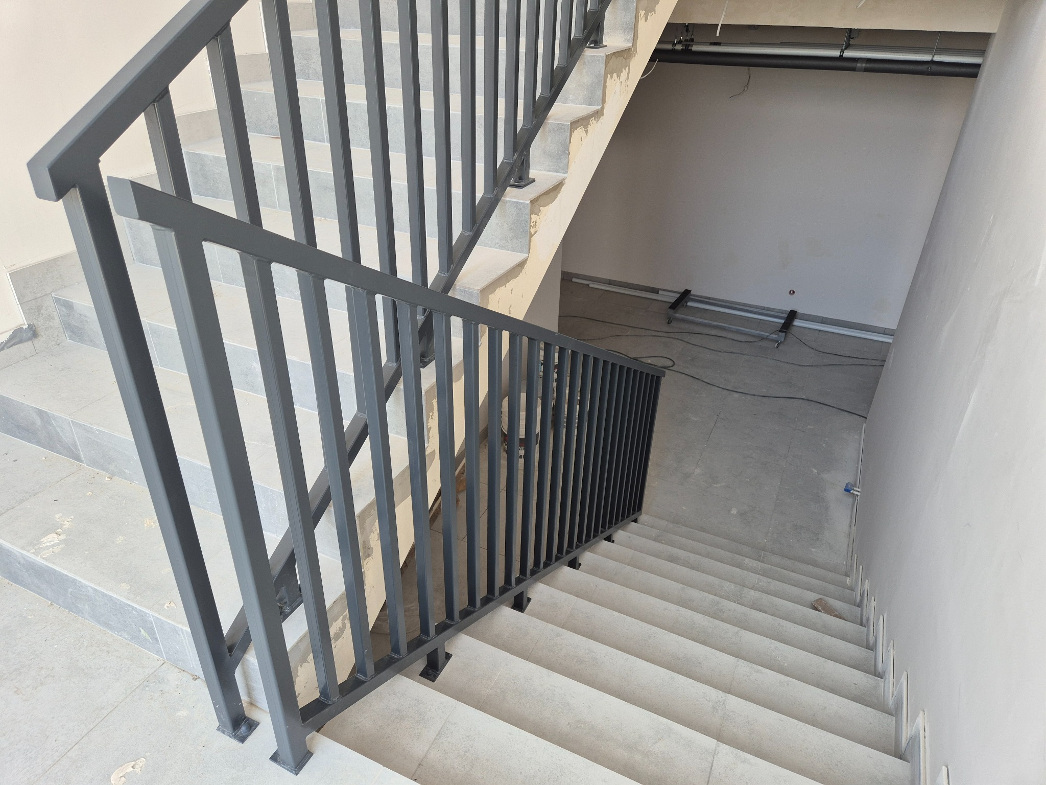 Wewnętrzne schody z szarymi stopniami i prostą, grafitową balustradą metalową, widok z góry na dół, w surowym stanie wykończenia.