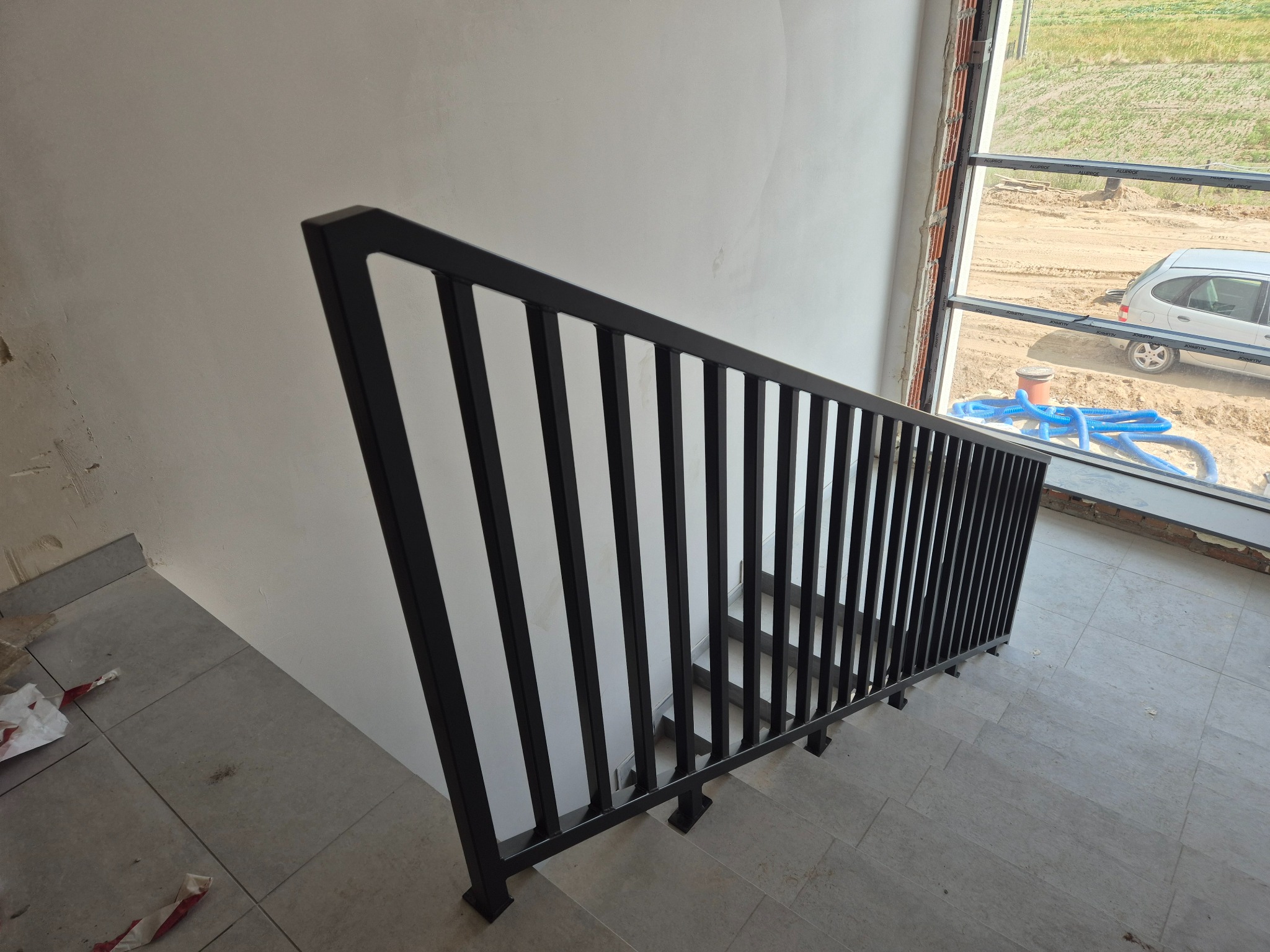 Czarna, metalowa balustrada schodowa o prostej konstrukcji, zamontowana w surowym wnętrzu z widokiem na plac budowy przez okno.